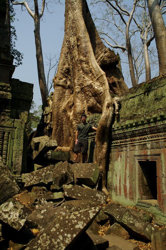 Justin standing on some fallen stones next to a tree on the roof of Ta Prohm in Angkor, Cambodia