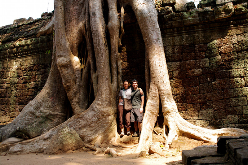 Stephanie and Justin standing between the humongous roots of tree on the wall of Ta Prohm in Angkor, Cambodia