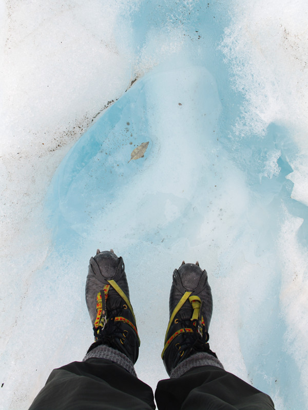 Looking down at my boots and crampons against some blue ice on Franz Josef Glacier